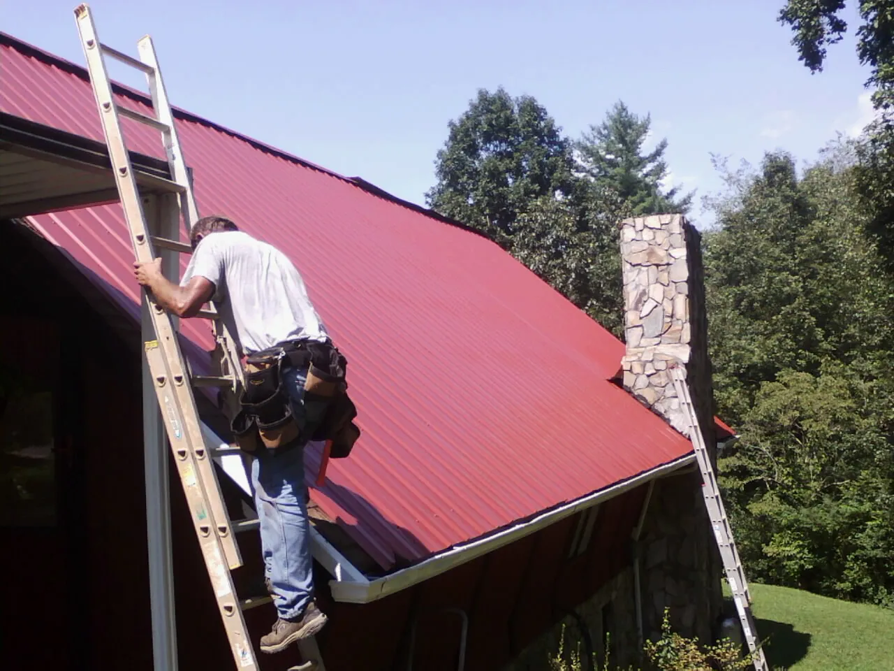 Roofer on a ladder beside a red metal roof and stone chimney — Soffit & Fascia Repair by Roofing Brothers in Worcester